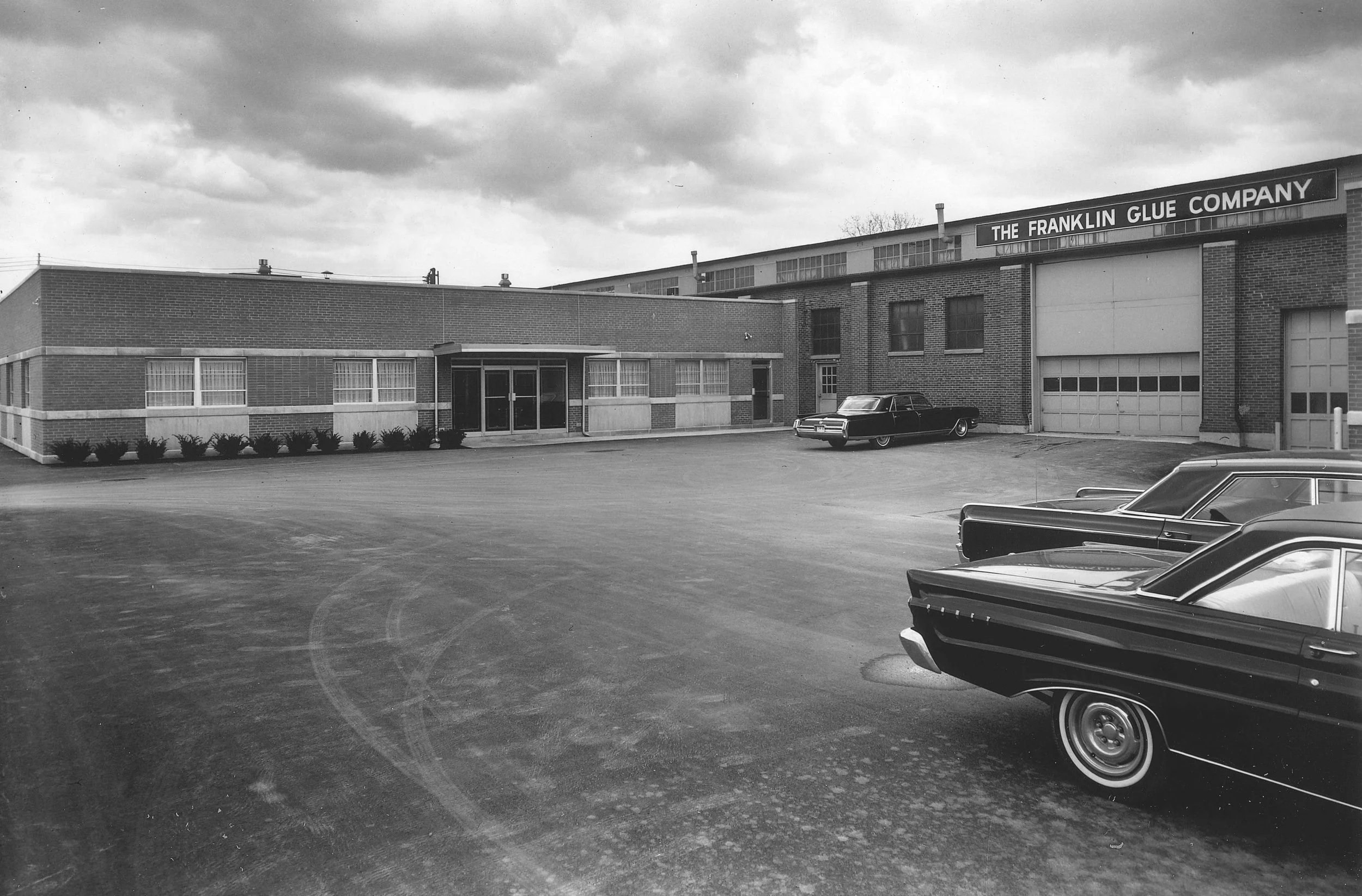 Black and white photo of a commercial building and several vintage cars parked in the lot.